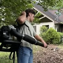 A man holding a leaf blower outdoors near a house, with trees and a garden in the background.
