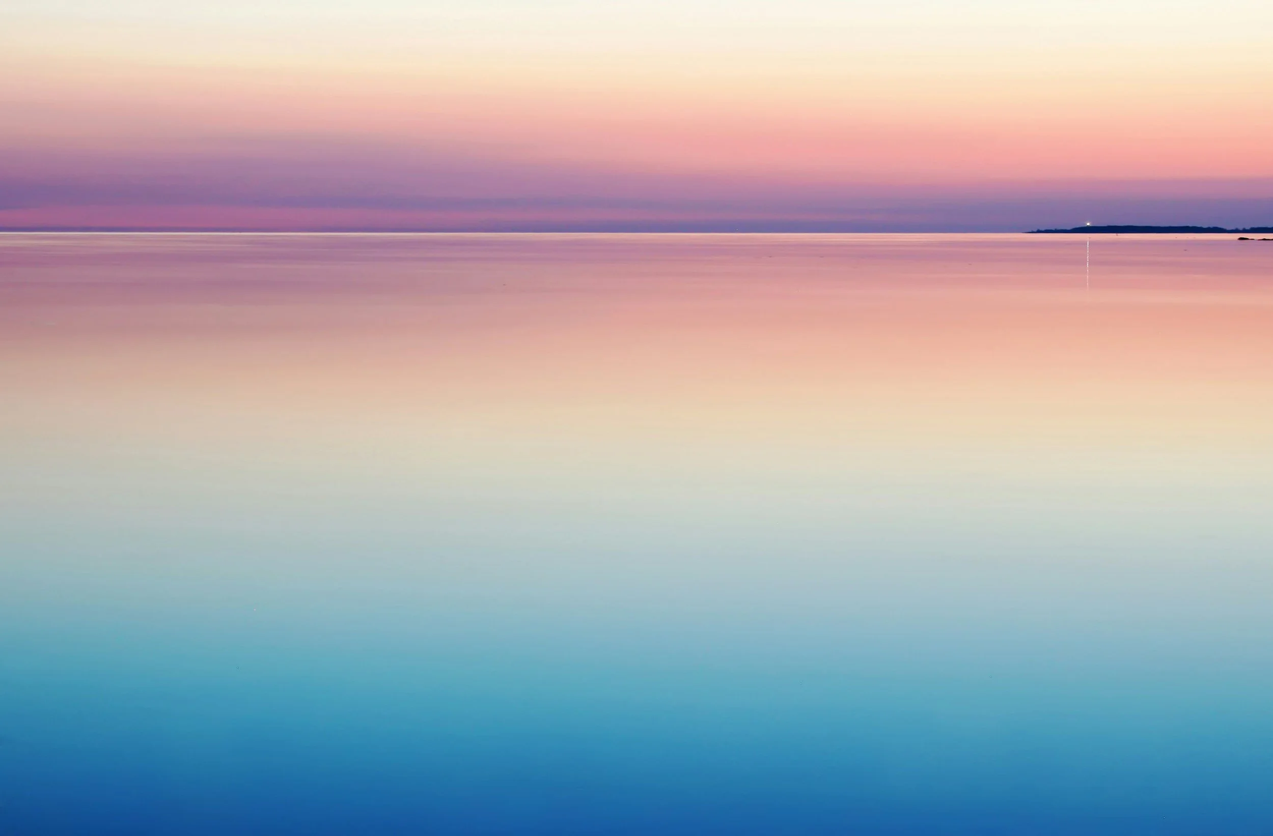 A serene scene of a calm body of water reflecting a pastel-colored sky at sunset, with a distant landmass on the horizon.