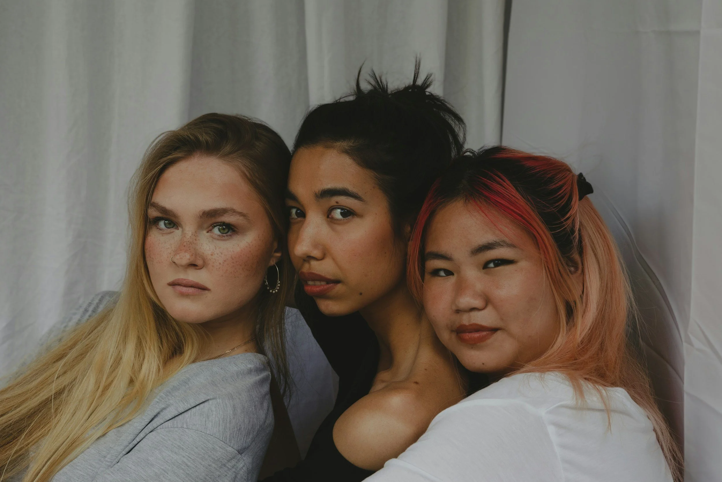 Close-up of three young women posing closely together in front of a light-colored curtain. The woman on the left has long blonde hair and freckles, the woman in the middle has short black hair with a tuft standing up, and the woman on the right has shoulder-length red hair with black streaks.