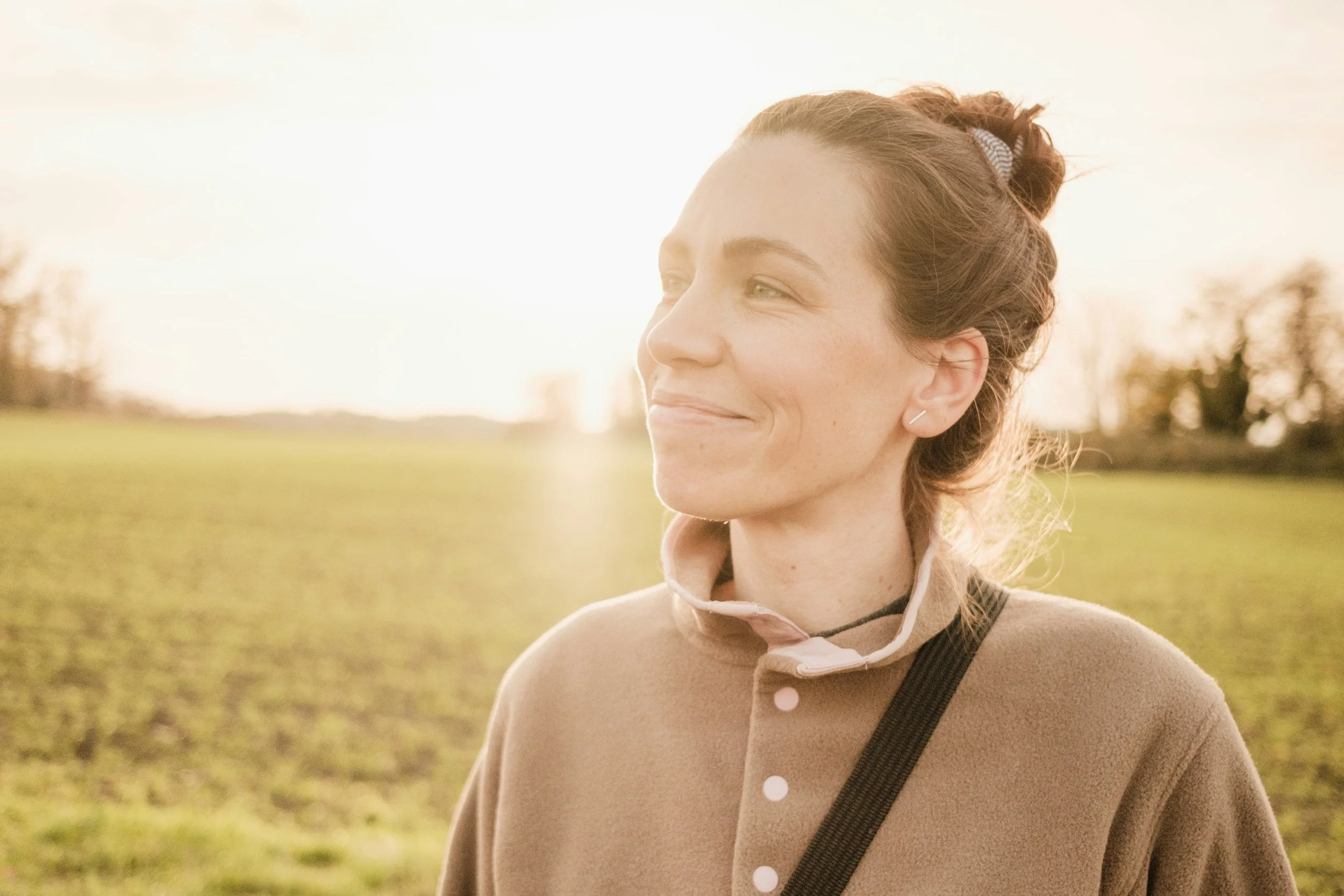 A woman with red hair tied in a bun and light skin smiling outdoors during sunset, wearing a beige coat and a black strap across her chest.