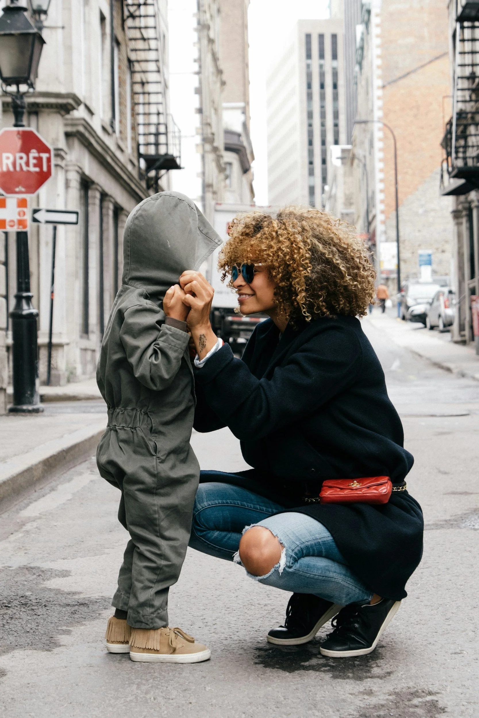 A woman crouching down, smiling, and holding hands with a young child on an urban street. The woman has curly hair and wears sunglasses, ripped jeans, a black jacket, and black sneakers. The child is dressed in a hooded jacket, beige fringed shoes, and is facing the woman with their hands held together.