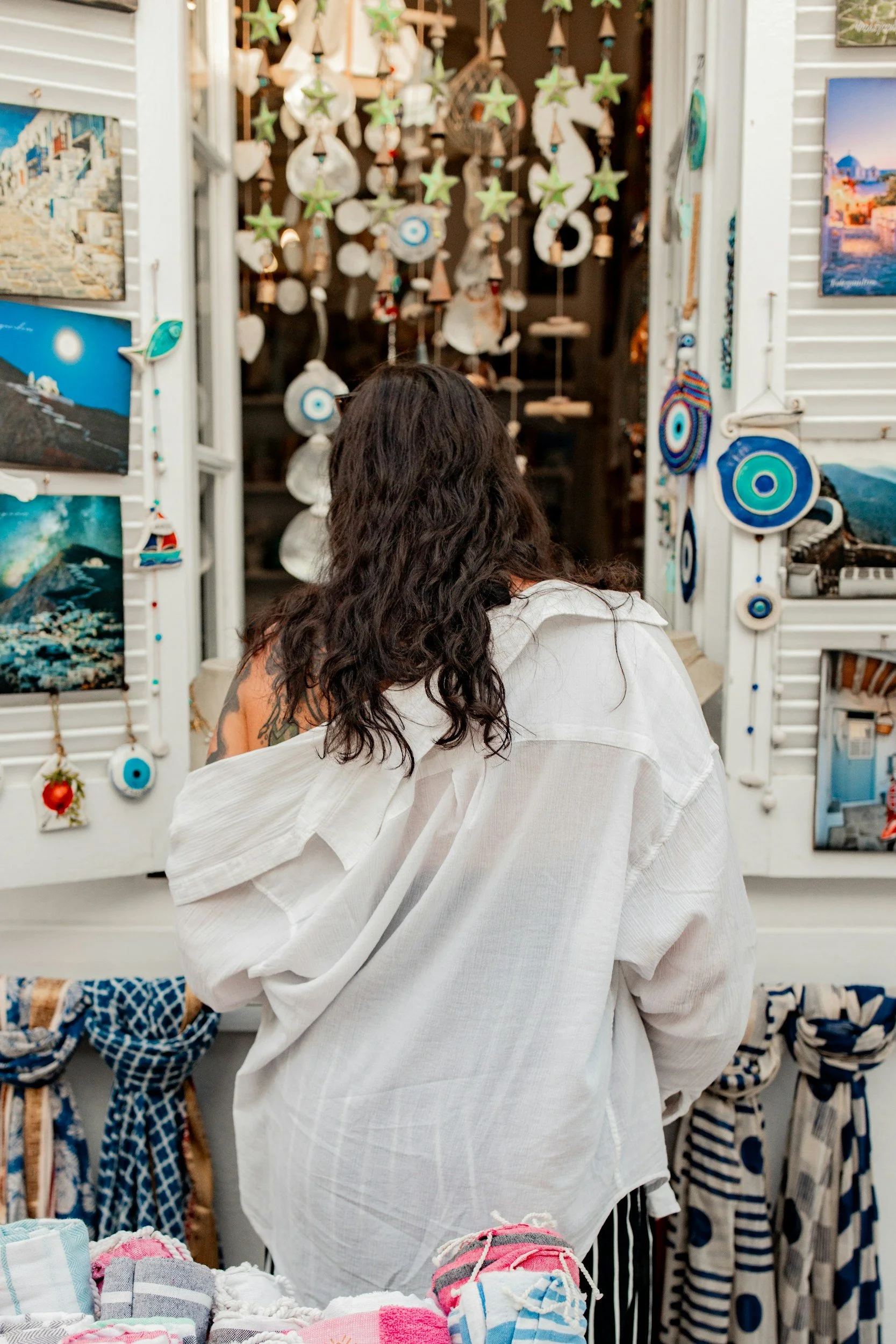 A person with long dark hair wearing a white shirt is looking at home decor items in a shop. The background displays hanging decorations and framed pictures, with additional clothing items on racks.