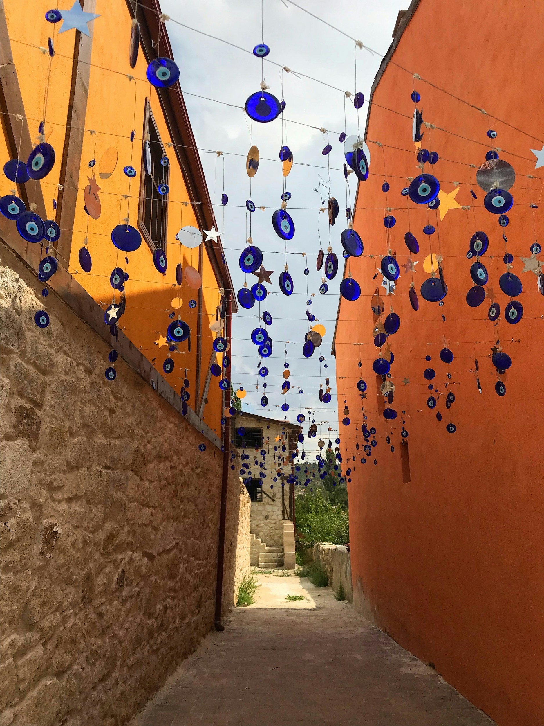 Narrow alleyway decorated with hanging blue evil eye charms and star-shaped ornaments, flanked by rustic stone and orange walls, with a stone staircase at the end.
