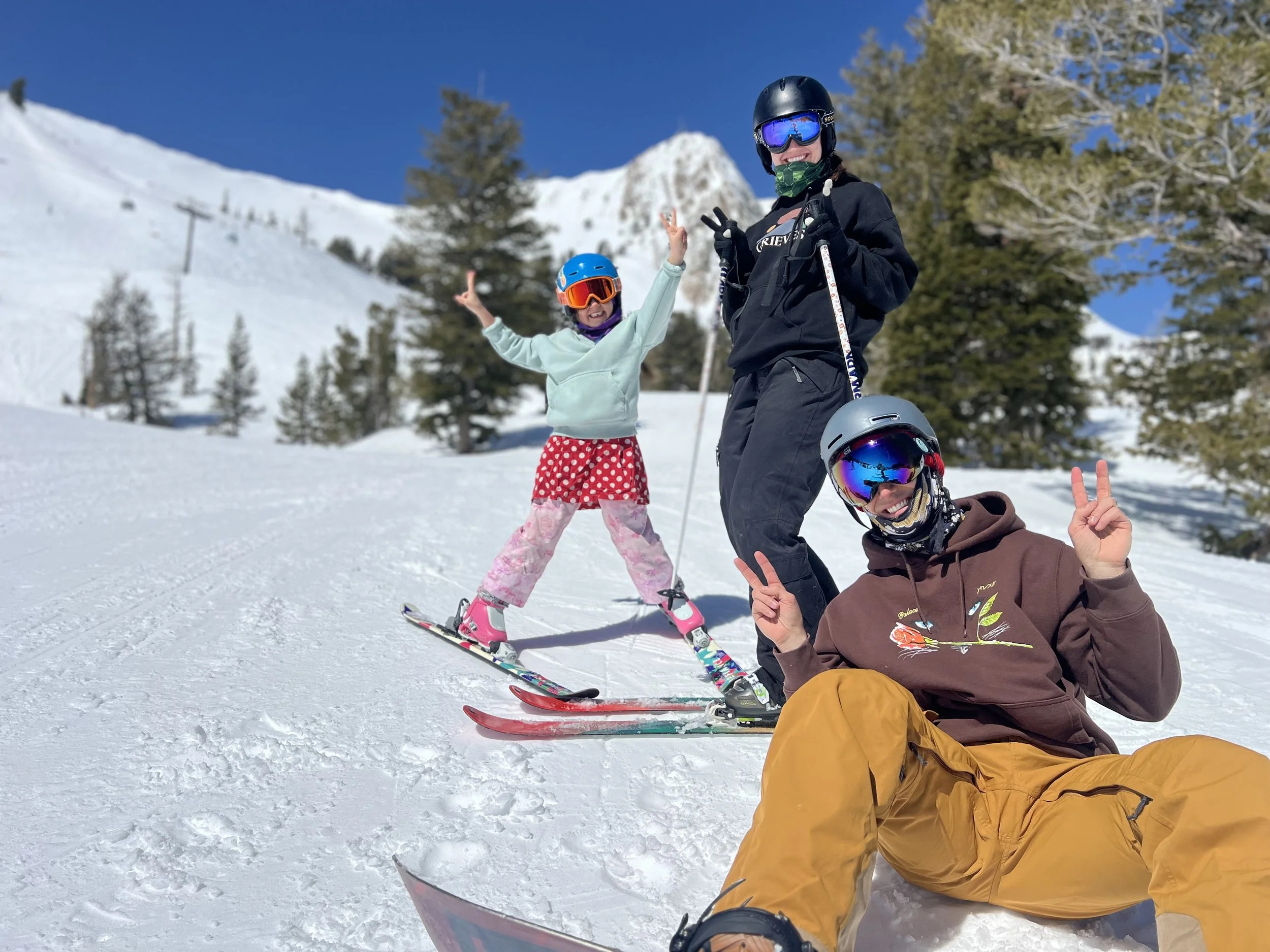Three people on a snowy mountain, wearing ski gear and helmets, with trees and snow-covered peaks in the background, smiling and making peace signs.