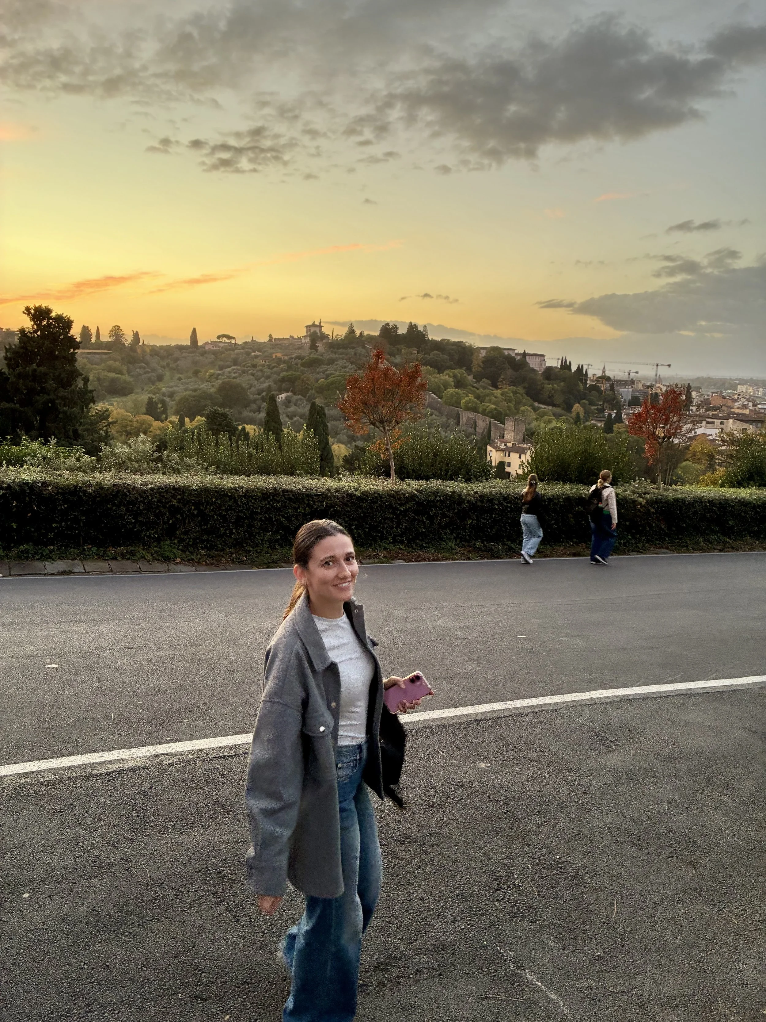 A young woman wearing a gray jacket, white shirt, and blue jeans walking along a road at sunset. She smiles at the camera and holds a pink phone. In the background, two other people walk along the same road, with trees, hills, and a cityscape in the distance under a partly cloudy sky.