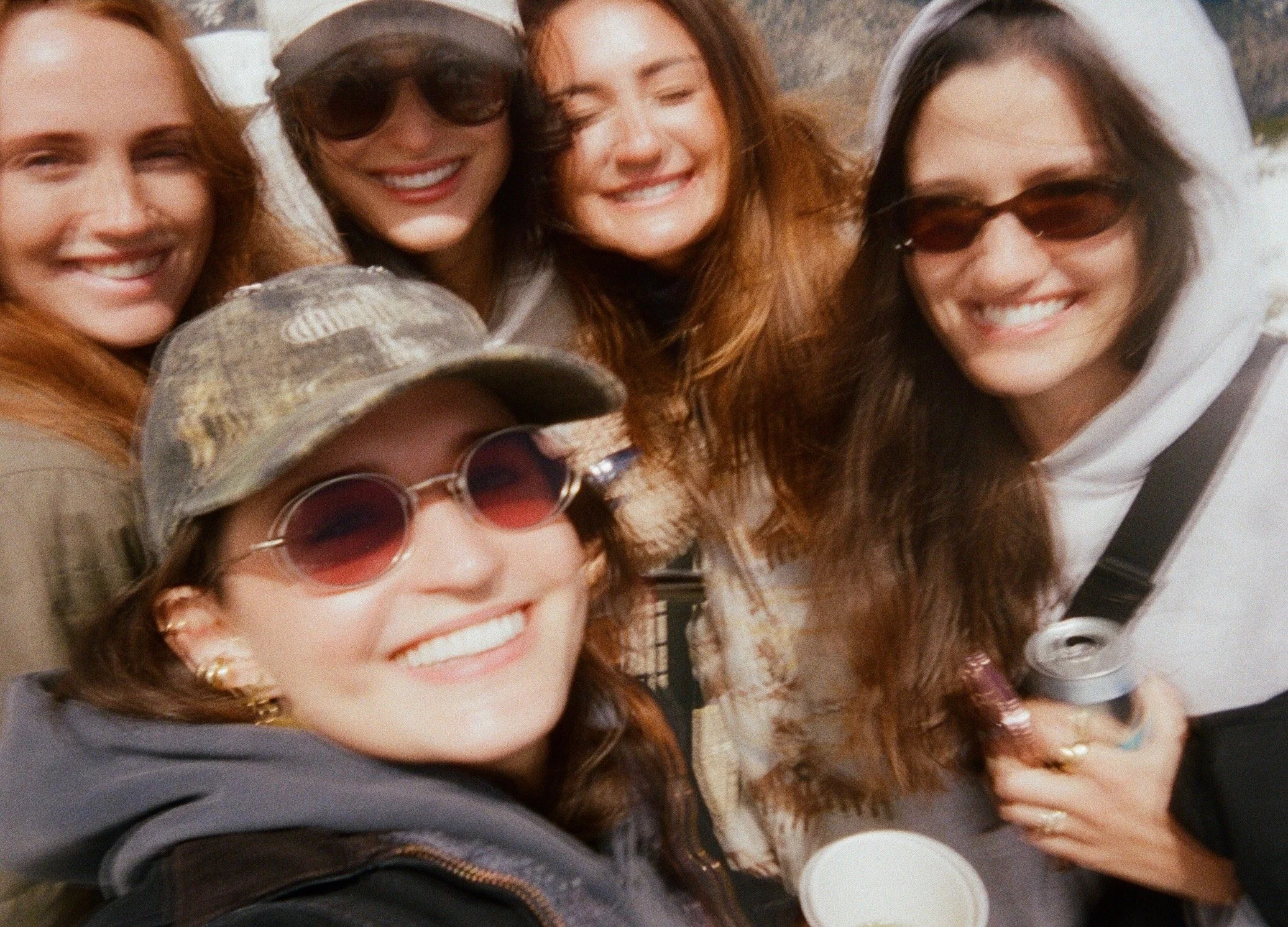 A group of five women smiling and taking a selfie together, wearing casual clothing and sunglasses, with one woman in a camouflage hat.