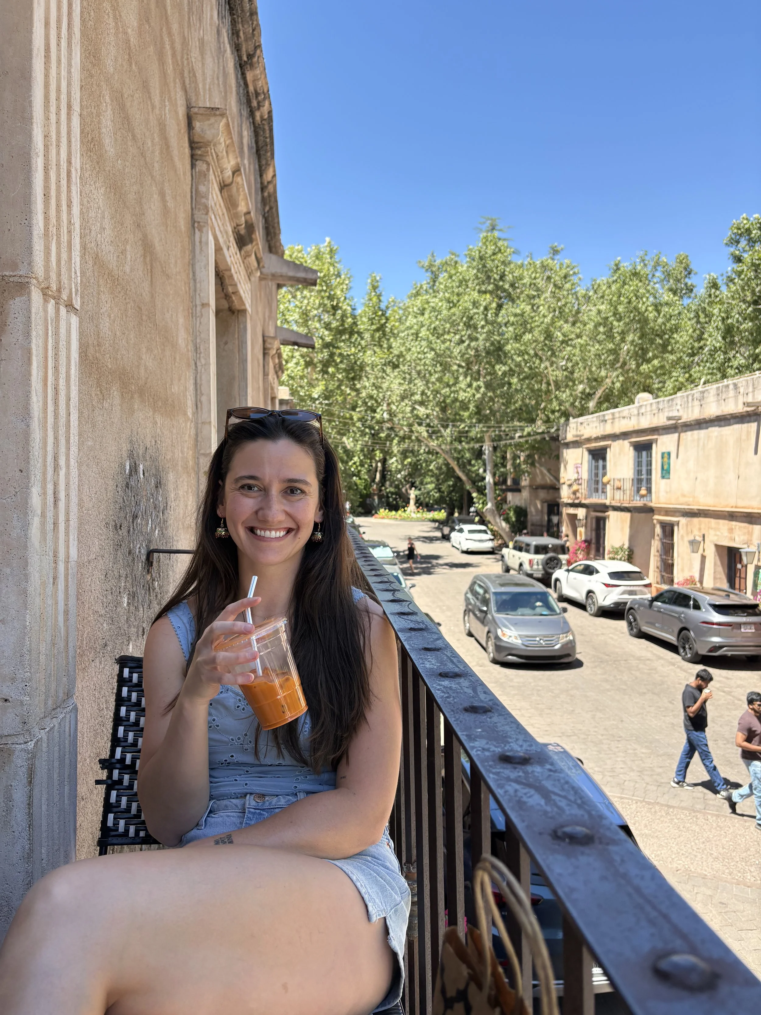 A young woman sitting on a balcony holding a plastic cup with orange beverage and a straw, smiling at the camera. She has long dark hair, sunglasses on her head, and is wearing a blue sleeveless top and denim shorts. The background shows a street with parked cars, two people walking, a tree, and a historic building with beige walls under a clear blue sky.