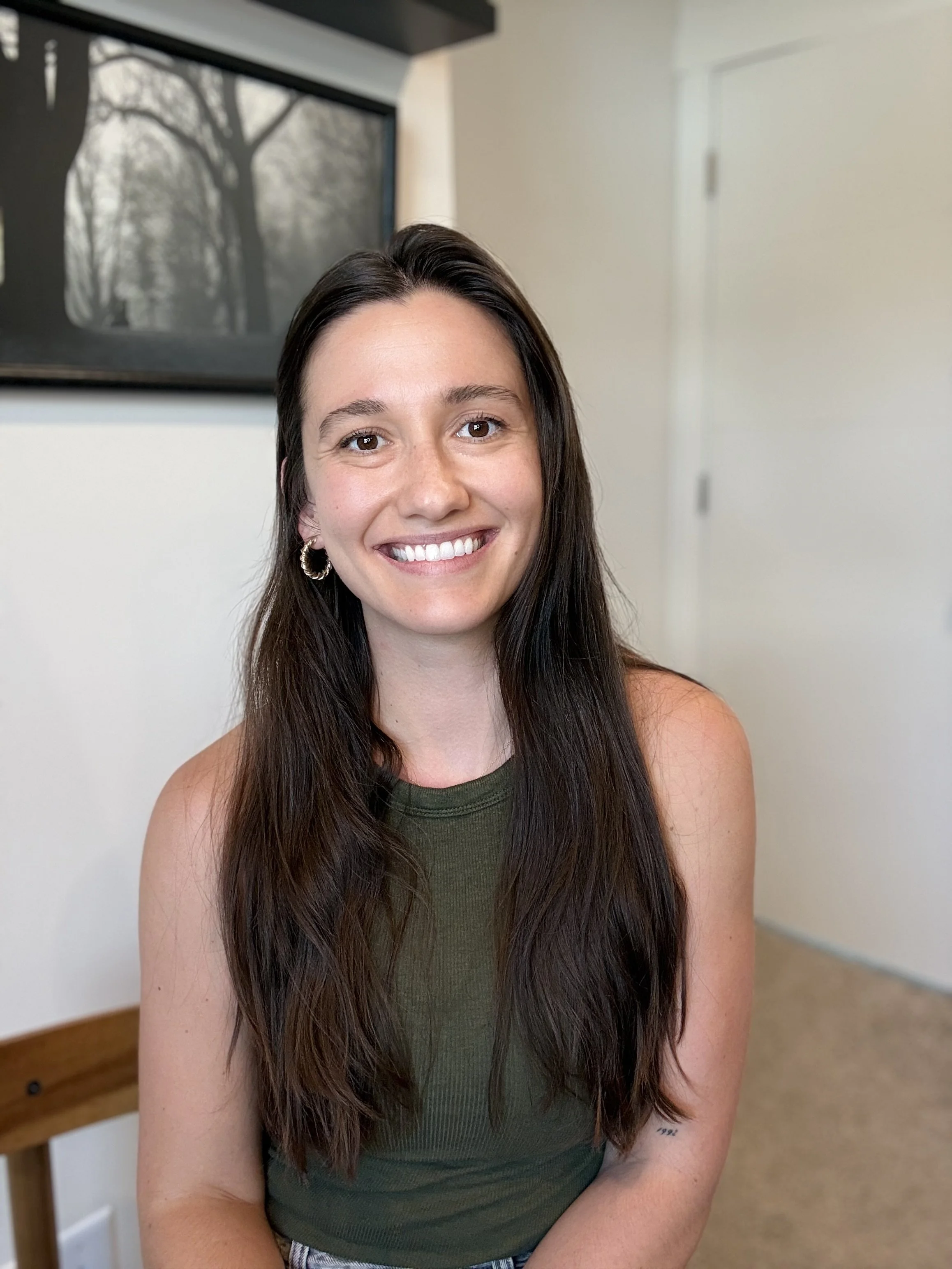 A woman with long dark hair smiling, wearing an olive green sleeveless top, sitting indoors with a framed black and white photo of trees on the wall behind her.