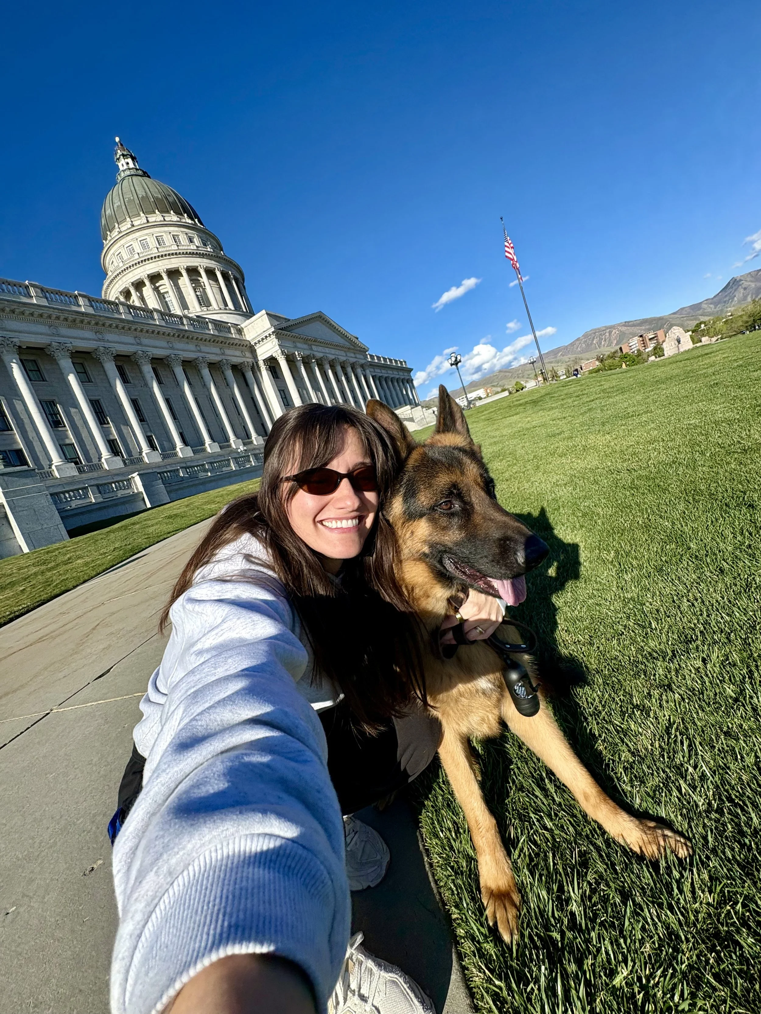 Woman taking a selfie with her dog in front of the Utah State Capitol building on a sunny day.