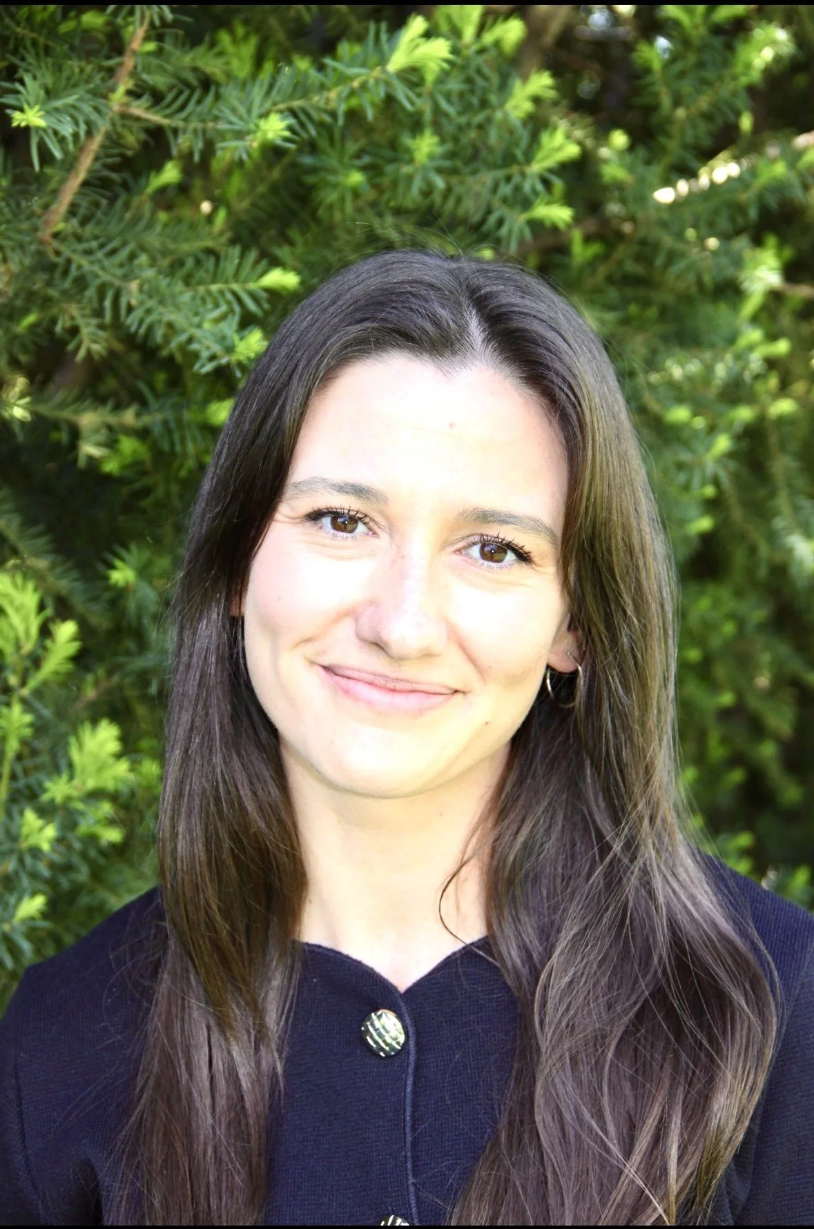 A woman with long brown hair and light skin smiling outdoors in front of green foliage.