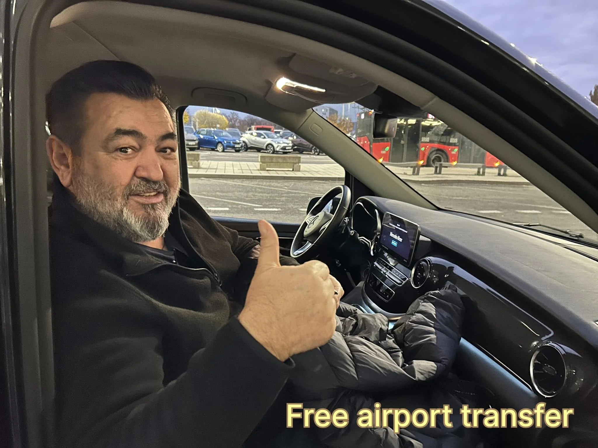 A man with a beard and dark hair giving a thumbs up inside a car at an airport, with a parking lot and a red bus visible through the window.
