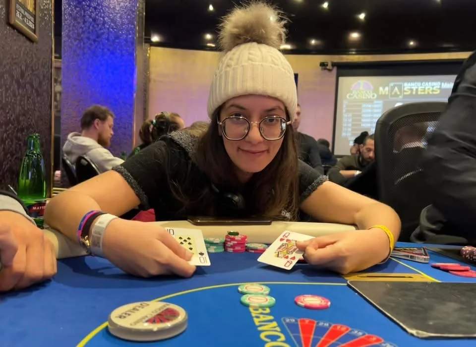 A young woman with glasses and a beige knit beanie with a pom-pom is sitting at a poker table in a casino, holding playing cards and smiling. There are poker chips, a smartphone, and drinks on the table, with other people in the background.
