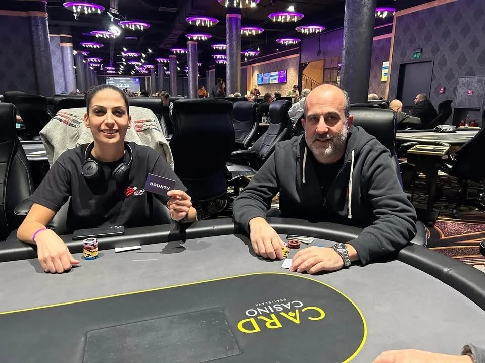 Two people seated at a poker table in a casino, with poker chips and cards, surrounded by other players and casino lights.