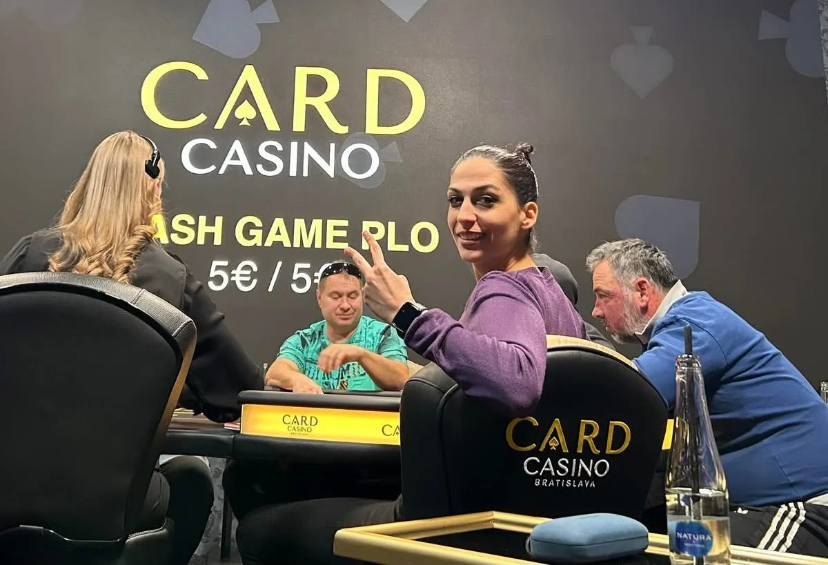 People playing poker at a casino table with a woman smiling and pointing at the camera, in front of a wall that says 'CARD CASINO' and 'CASH GAME PLO 5€ / 5€'.