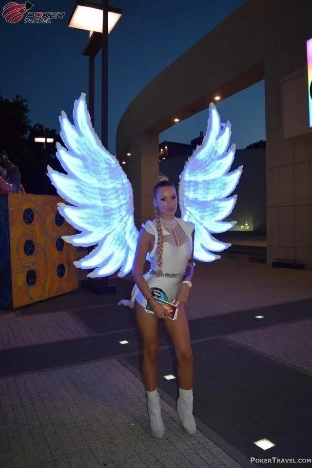 A woman dressed as an angel with large, illuminated white wings, standing outdoors at night near a building with bright lights.