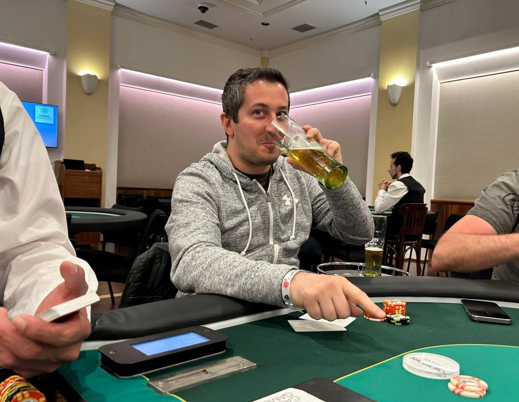 A man with short dark hair and a gray hoodie drinking beer at a poker table in a casino conference room, with poker chips, cards, and a small electronic device on the table.