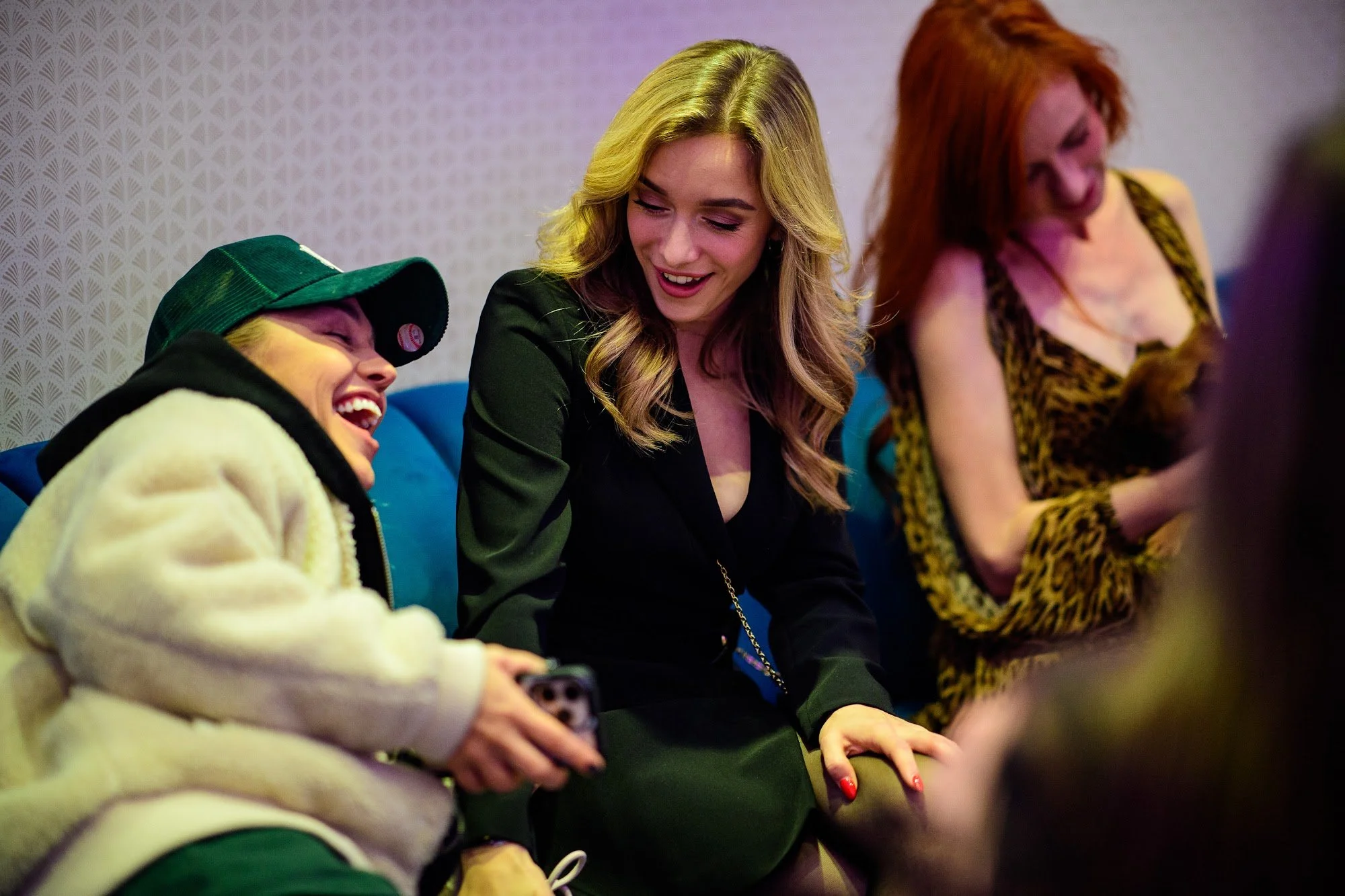 Four people sitting on a bench, smiling and talking. The person on the far left is wearing a green cap and cream-colored jacket. The second person is a blonde woman in a black blazer. The third person is a woman with red hair in a leopard print top. 