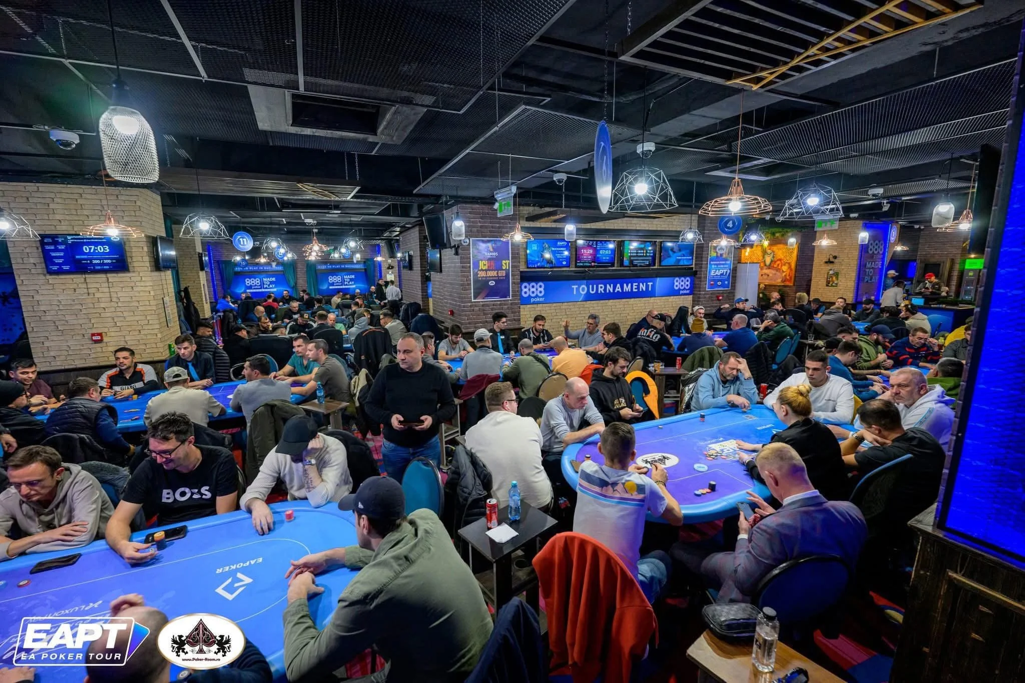 Several people playing poker at blue tables in a brightly lit indoor casino or event space, with digital screens and tournament signage in the background.