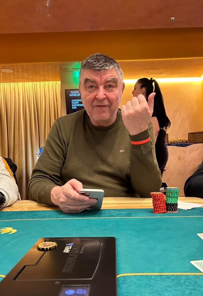 A man sitting at a poker table holding a smartphone, with poker chips in front of him, in a casino setting.
