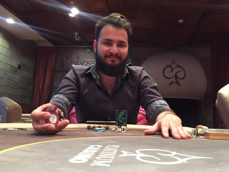 A man with dark hair and a beard sitting at a poker table with poker chips, cards, and a small stack of green chips in front of him, in a room with wood-paneled walls and a decorative card suit symbol in the background.