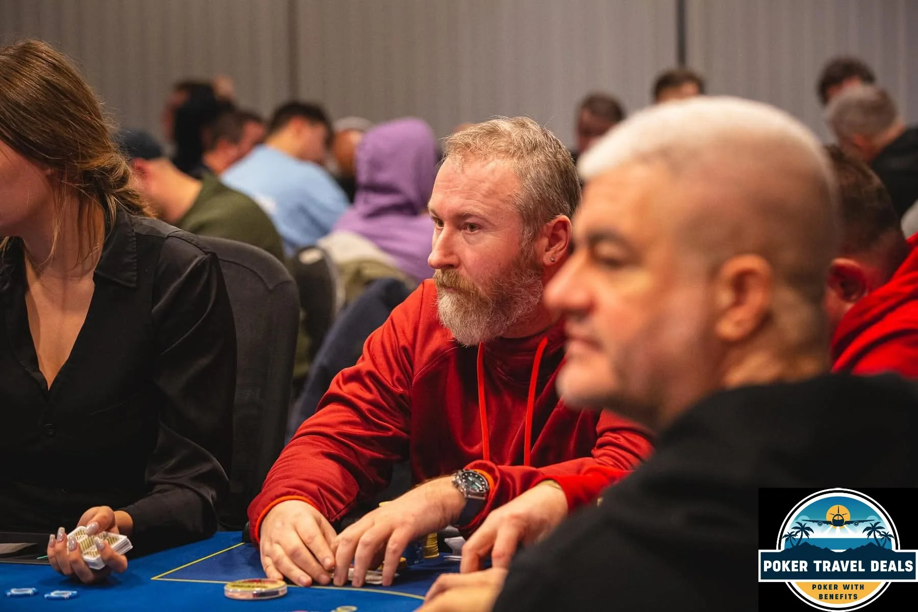 A group of people sitting at a poker table, playing cards and chips, with a background of other players and a poker tournament setting.