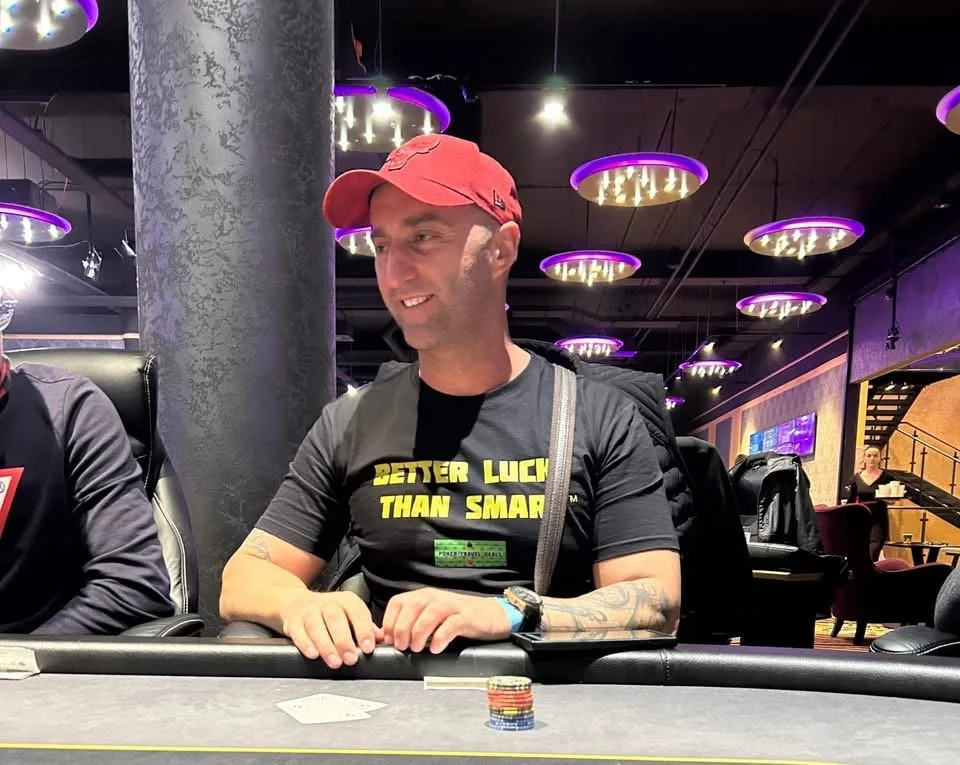 Man wearing a red cap and black T-shirt with yellow text, sitting at a poker table with poker chips in front of him, in a casino or poker room with purple lighting and modern decor.