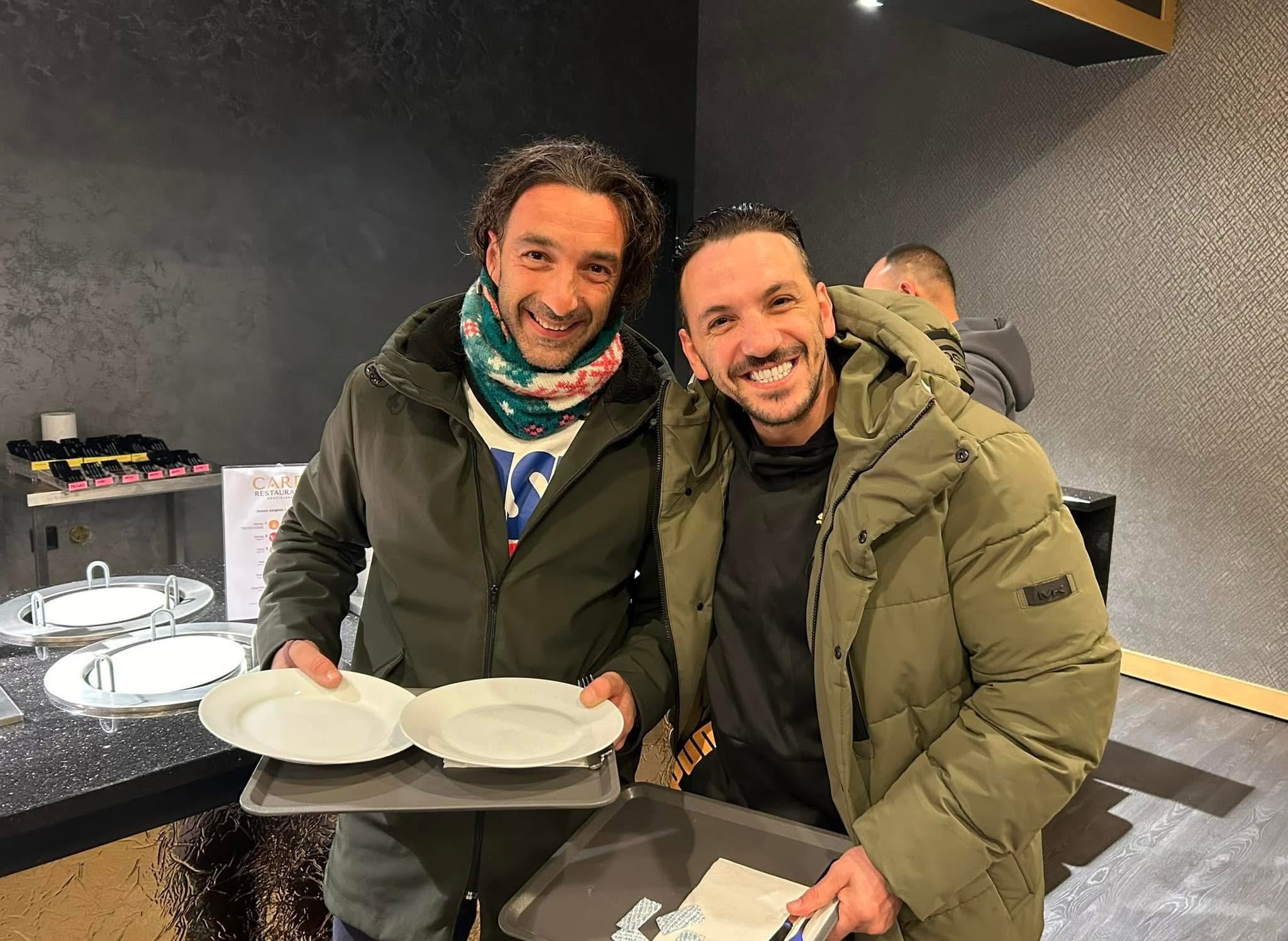 Two men smiling and posing at a dining area, holding empty white plates. One man has curly hair and a colorful scarf, and the other has short dark hair and a green jacket. Behind them, there is a man with a shaved head in the background, and the sett