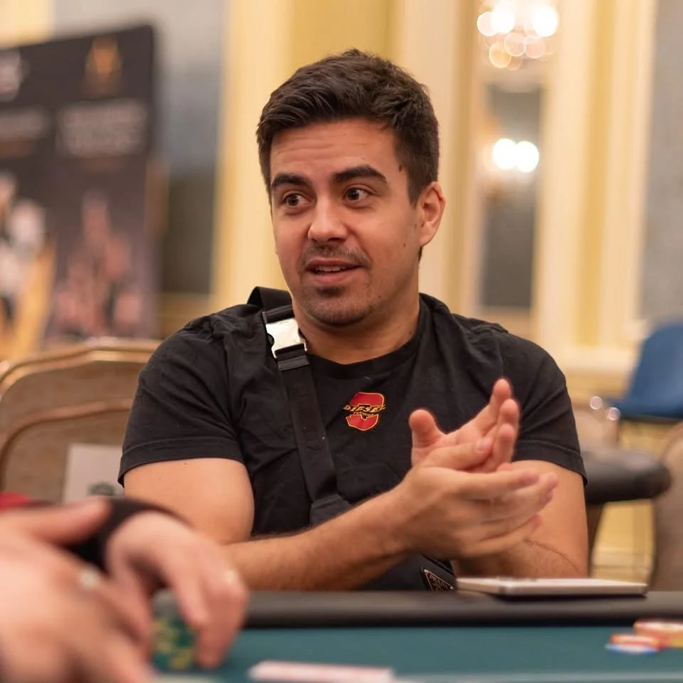 A man with dark hair and a short beard speaking and gesturing with his hands at a poker or casino table, with a blurred background of a luxurious room with yellow walls and large mirrors.