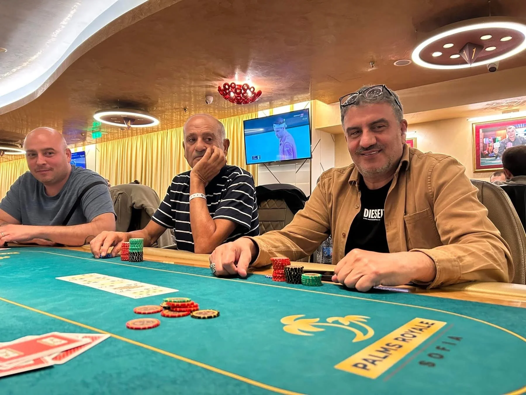 Three men sitting at a poker table with chips in front of them. The background features yellow curtains, a flat-screen TV, and decorative ceiling lights.