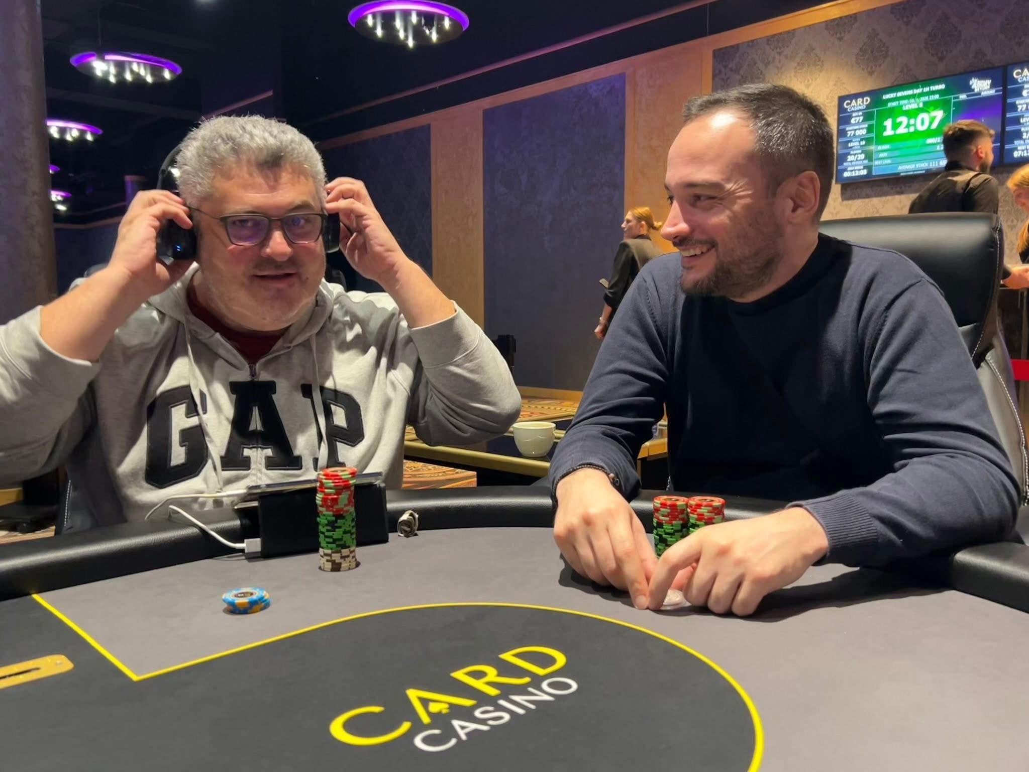Two men sitting at a poker table in a casino, smiling and playing poker with stacks of poker chips on the table. One man is wearing glasses and headphones, while the other is smiling and looking at the first man. A digital display in the background s