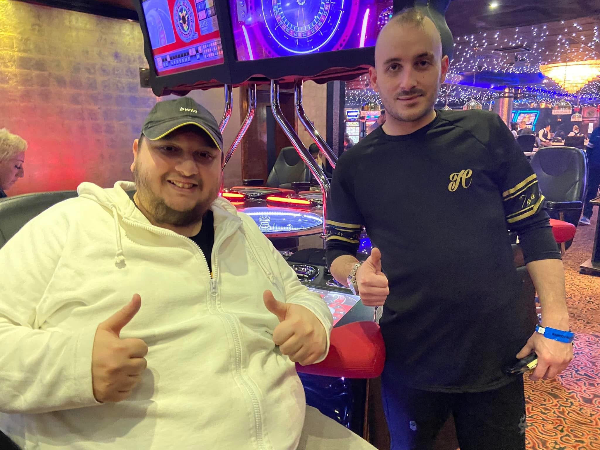 Two men sitting at a table in a casino, giving thumbs up and smiling. One is wearing a white hoodie and the other a black shirt with yellow accents. Slot machines and large screen displays are visible in the background, with colorful lights and a fes