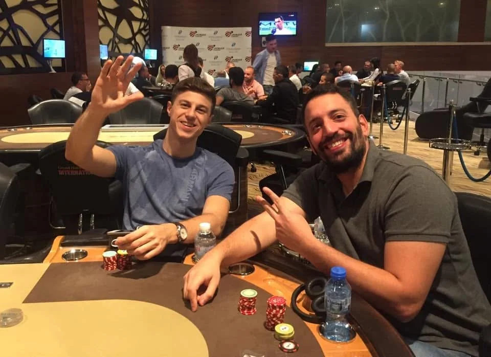 Two men sitting at a poker table, smiling and making gestures. There are poker chips, water bottles, and a phone on the table. In the background, multiple people are gathered around tables in a casino or poker room setting, with screens and a TV moun