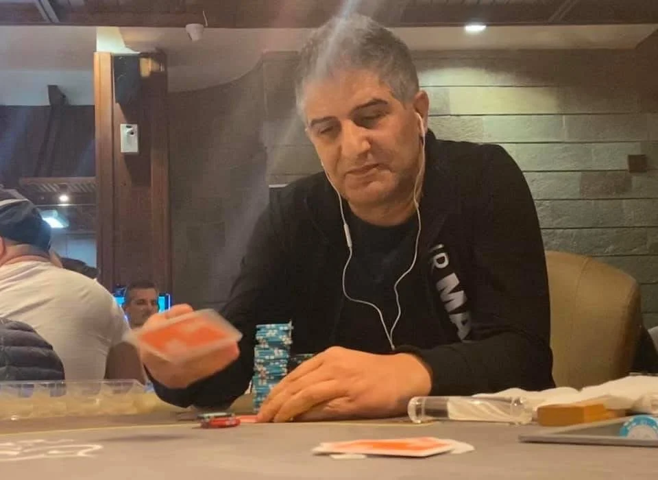 A man with gray hair is sitting at a poker table, stacking poker chips and holding a hand of cards. He is wearing a black long-sleeve shirt, has white earbuds, and appears focused.