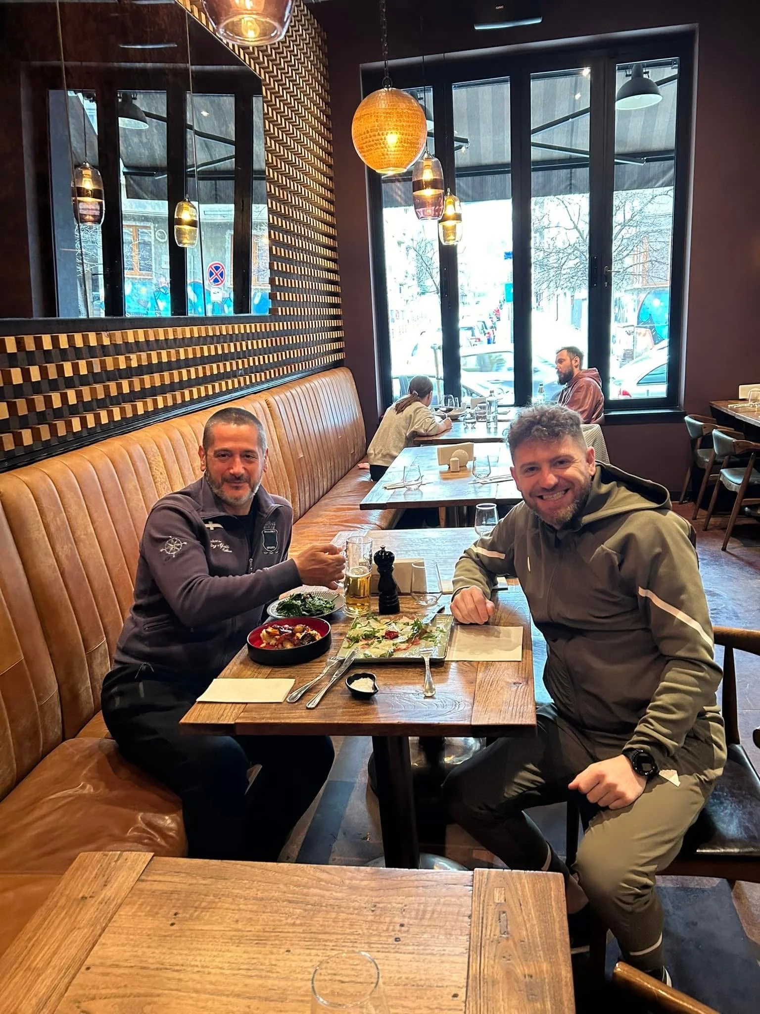 Two men sitting at a wooden table in a restaurant, smiling at the camera, with food and drinks in front of them. There are empty tables, large windows, hanging pendant lights, and a decorative wood detail on the wall.