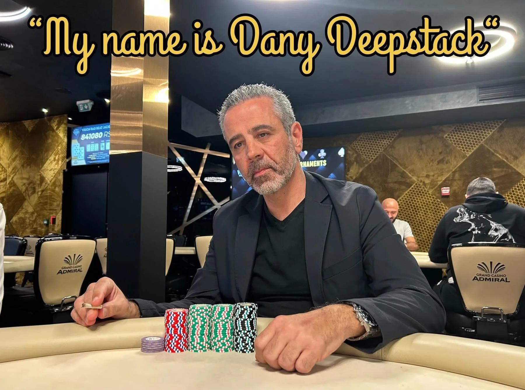 A middle-aged man with gray hair and a beard sitting at a poker table in a casino, with stacks of poker chips in front of him, wearing a black blazer and black shirt, with other casino patrons and gold-patterned walls in the background.