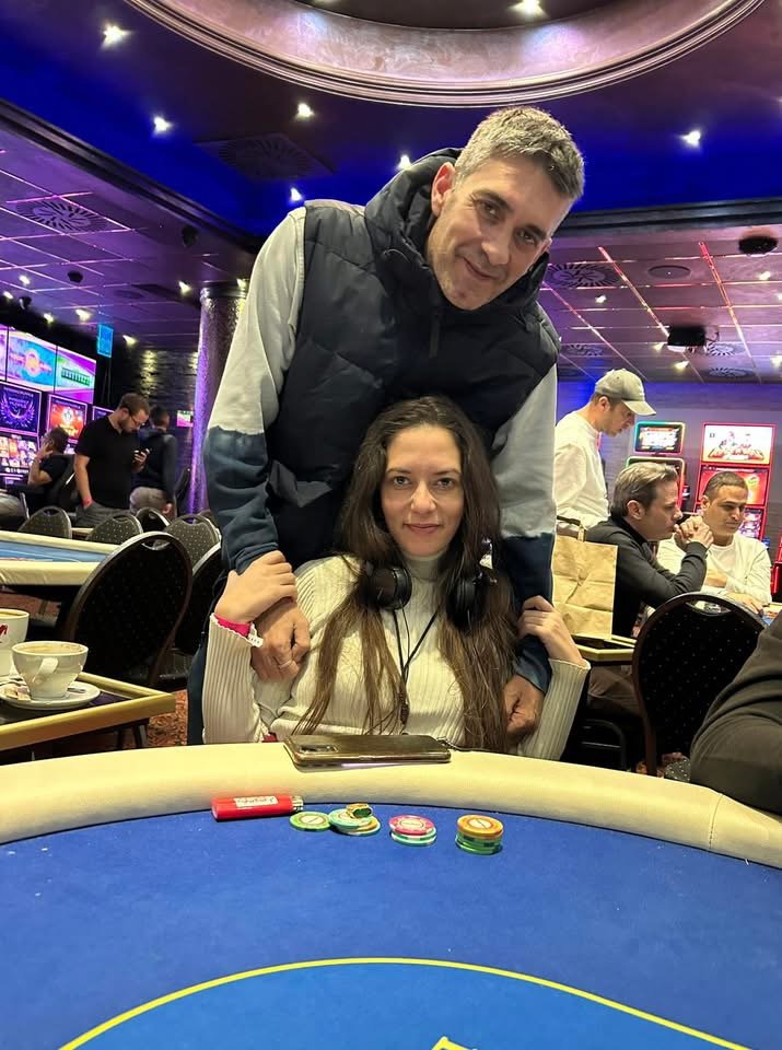 A man and woman at a casino table with poker chips, with people in the background playing slot machines.