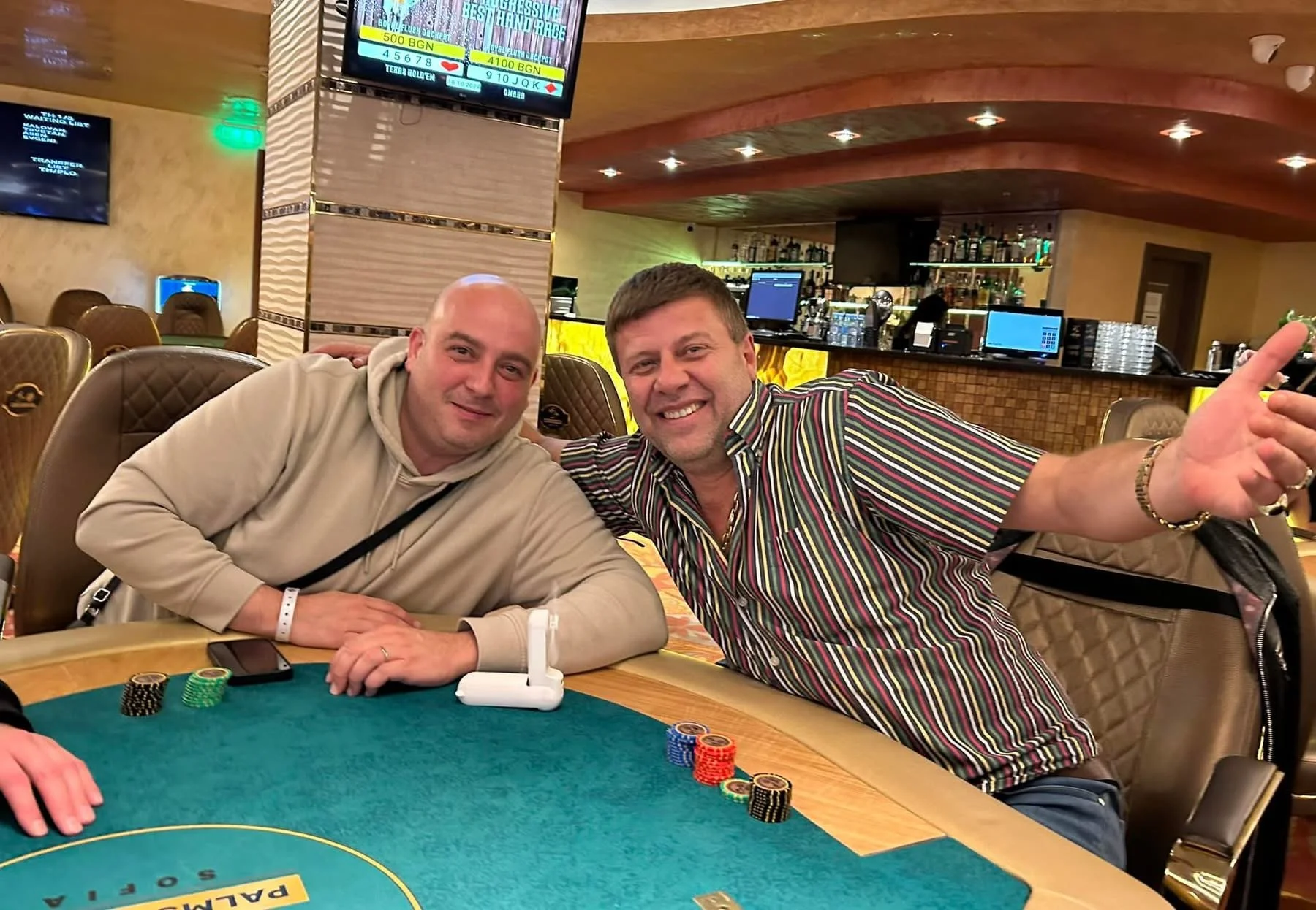 Two men sitting at a poker table in a casino, smiling at the camera. One is wearing a beige hoodie and the other a colorful striped shirt. Poker chips and a smartphone are on the table, and the background shows a bar and multiple electronic screens.