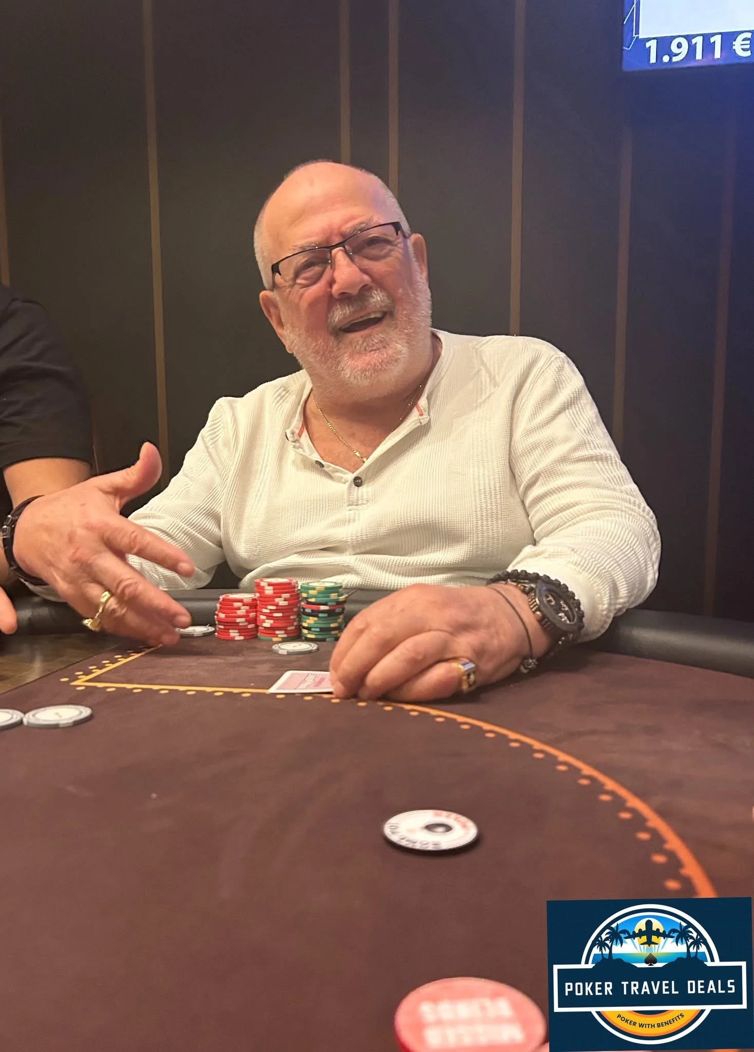 A man with glasses, white hair, and a beard smiling and gesturing with his hand at a poker table with poker chips. There is a poker travel deals logo in the bottom right corner.