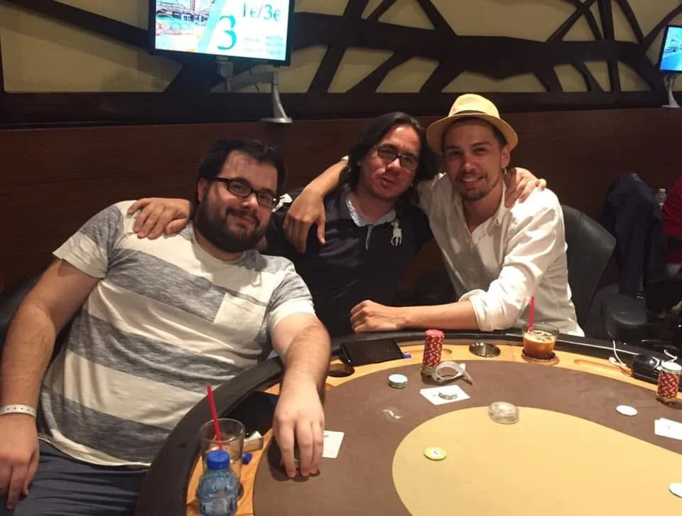 Three men sitting at a poker table in a casino, smiling and posing for the photo, with chips and drinks on the table.
