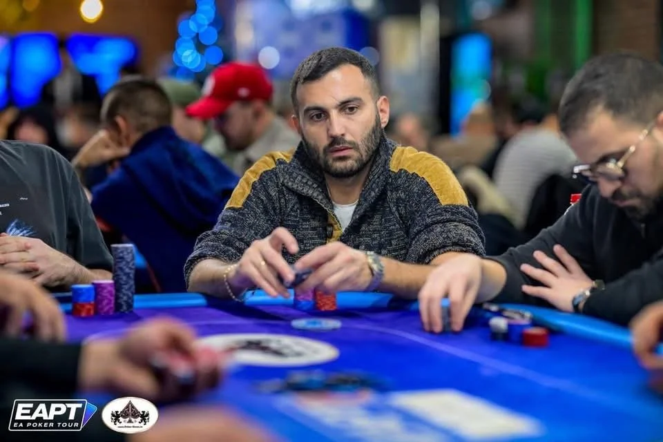 A man with short dark hair and a beard, wearing a dark sweater with yellow shoulder patches, is sitting at a poker table, holding poker chips. There are other players around the table, with poker chips and cards, in a lively casino or poker room envi
