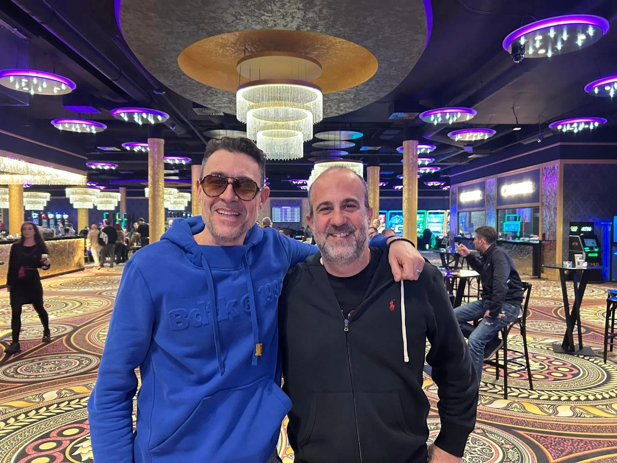 Two men smiling and posing for a photo in a brightly lit casino with colorful chandeliers and slot machines in the background.