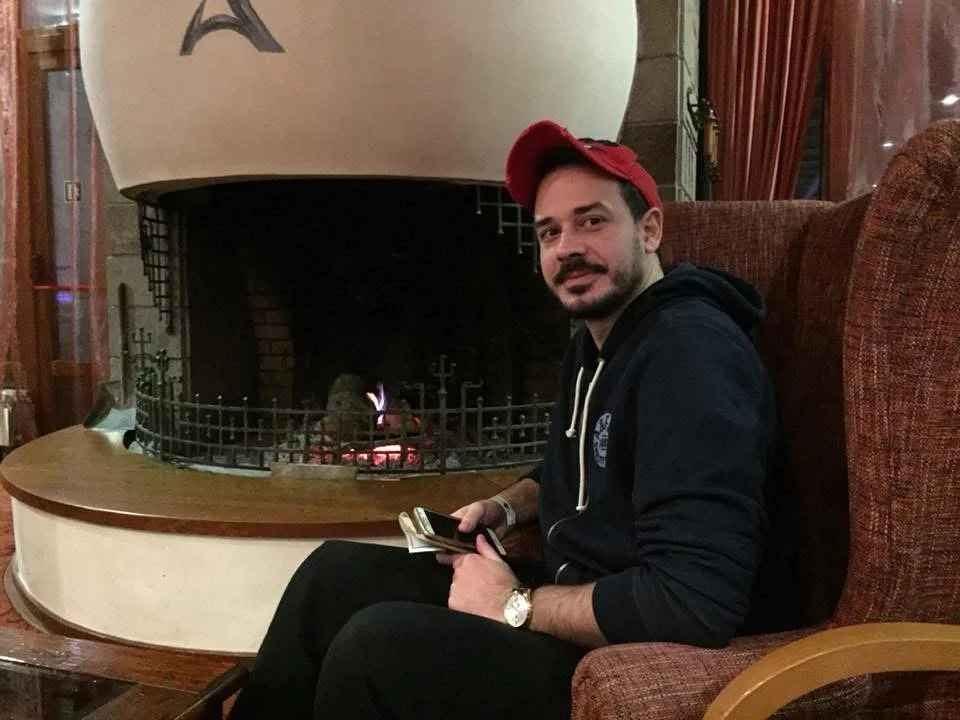 A man sitting in a red upholstered armchair by a fireplace, holding a wallet and a phone, wearing a black hoodie, a red cap, and a watch.