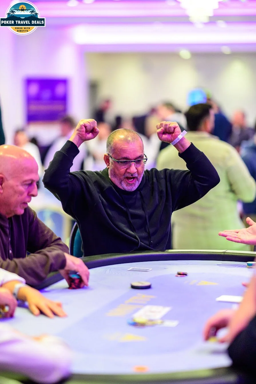 A man in glasses and black clothing celebrating at a poker table with arms raised and fists clenched, with other players and blurred background in a casino setting.