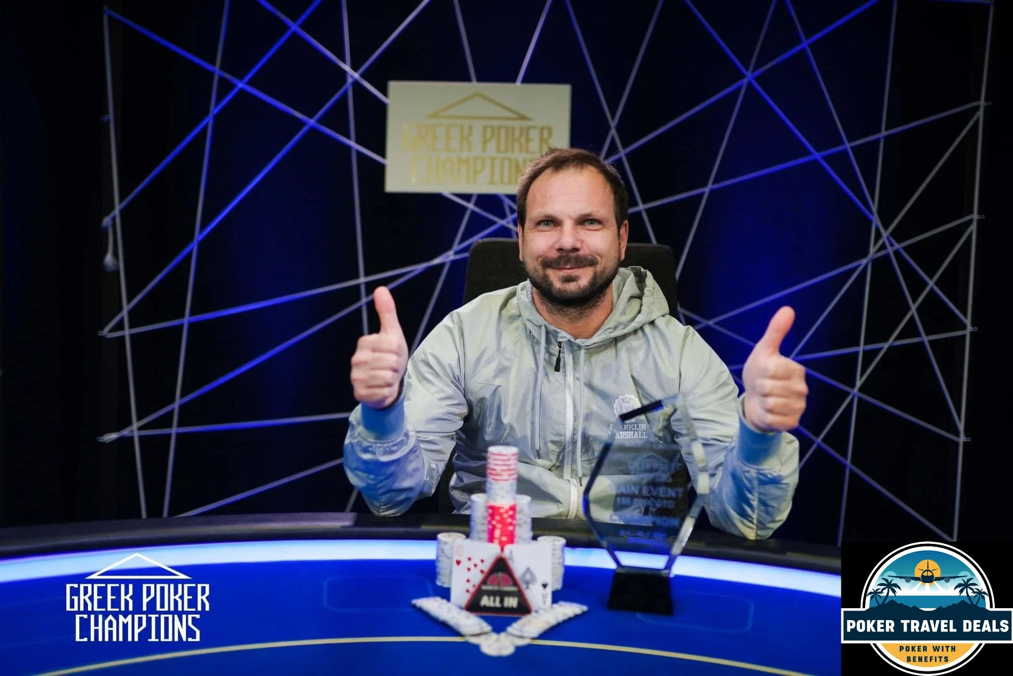 A man with a beard, sitting at a poker table, smiling and giving two thumbs up. The poker table has poker chips and playing cards, and there is a sign in the background that says 