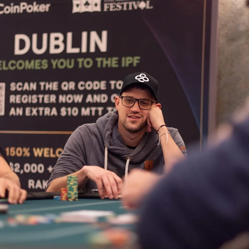 A man wearing glasses and a black cap sits at a poker table with poker chips stacked in front of him. He is resting his head on his left hand and smiling. There is a large sign behind him with text about the International Poker Festival in Dublin.