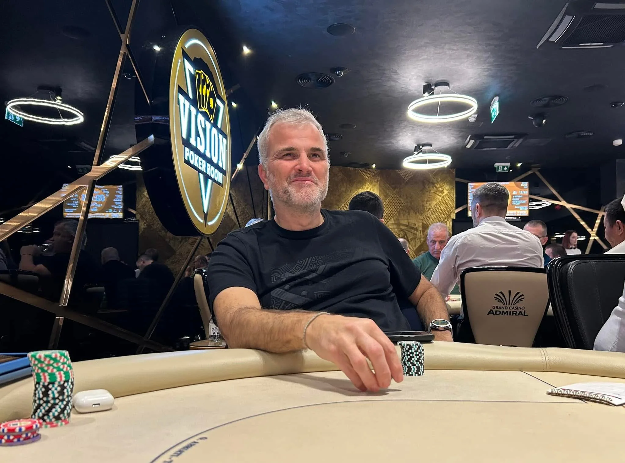 A man with gray hair and a beard playing poker at a casino table surrounded by other players. There are stacks of poker chips in front of him, and a sign in the background reads 'VISA POKER ROOM'. The casino's interior is decorated with dark colors a