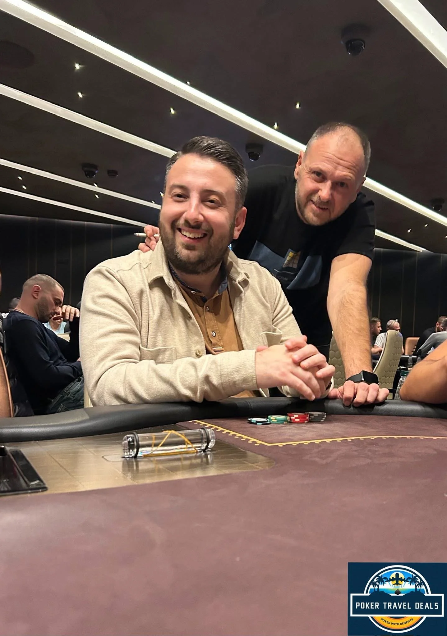 Two men smiling at a poker table with poker chips, one sitting and one standing, in a casino setting with other players in the background.