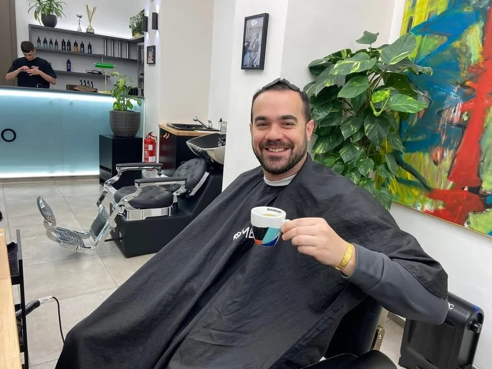 A man sitting in a barber's chair at a barbershop, smiling and holding a cup of coffee. He is wearing a barber cape. There are plants and colorful artwork on the wall behind him, and a barber station with tools in the background.