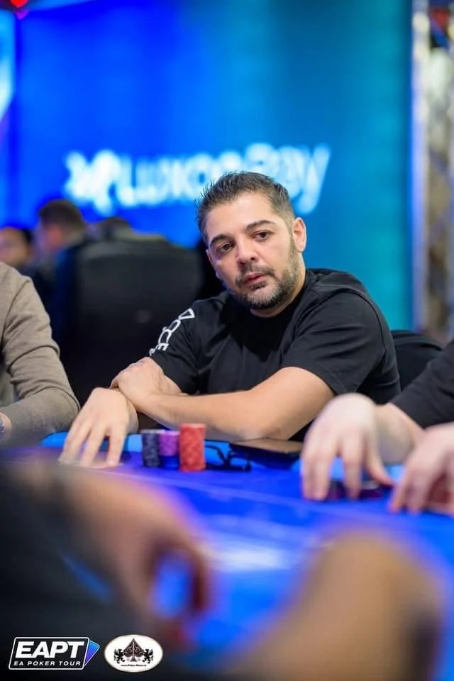 A man with dark hair and a beard playing poker at a poker tournament table with poker chips in front of him, focused on the game, in a casino or poker room environment.
