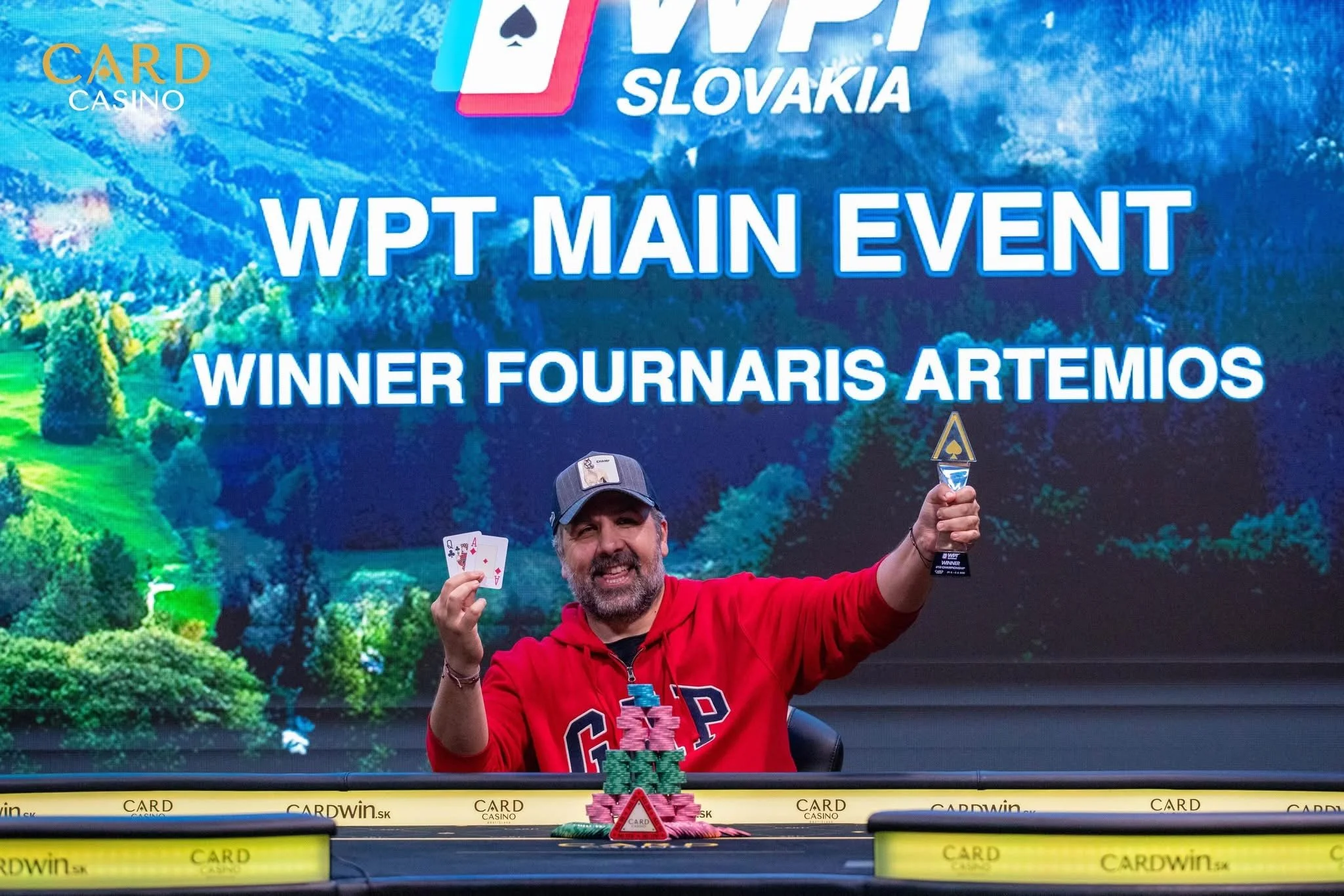 A man in a red shirt is celebrating after winning a poker tournament, holding a winning hand of two aces and a trophy. Behind him, a large screen displays the words 'WPT Main Event' and 'Winner Fournaris Artemios,' and a decorative pile of poker chip